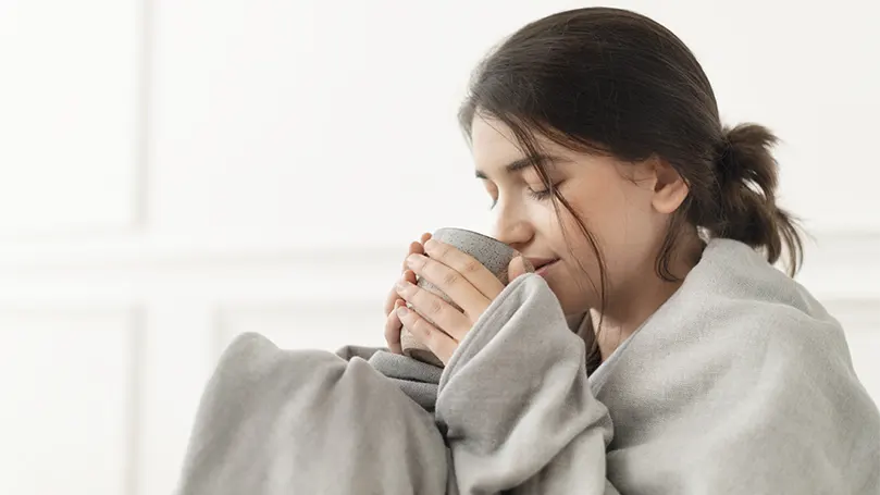 Een afbeelding van een vrouw die thee drinkt, gewikkeld in een verzwaarde deken voor diepe drukstimulatie.