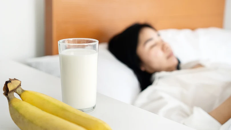 Een afbeelding van een slapende vrouw naast een nachtkastje met melk en bananen erop.