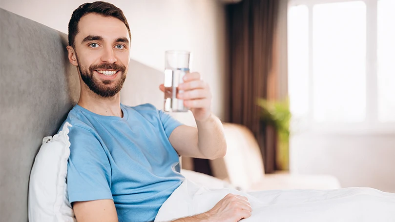 Een afbeelding van een man die een glas water drinkt in zijn bed voor bedtijd.