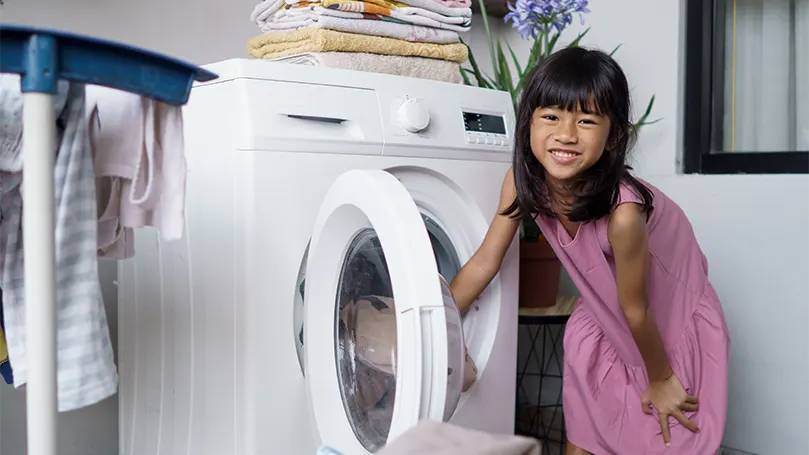 Een afbeelding van een klein meisje dat een deken in een wasmachine stopt.