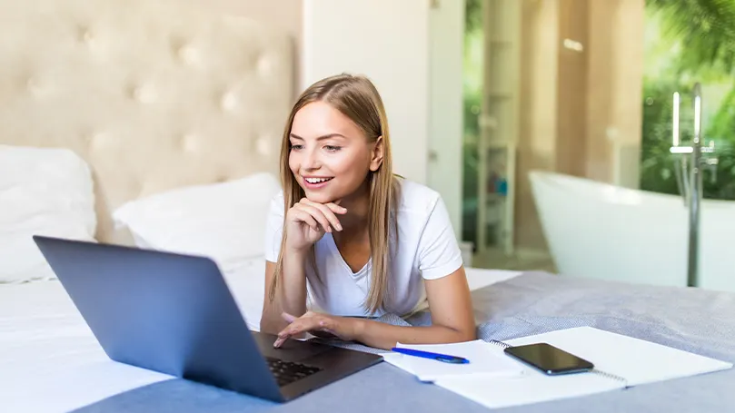 Een afbeelding van een vrouw die op haar bed ligt en op haar laptop werkt