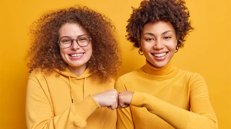 Een afbeelding van twee vrouwen in oranje truien die met elkaar op de vuist gaan en elkaar steunen