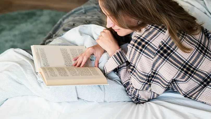 Een afbeelding van een vrouw die een boek leest in bed.